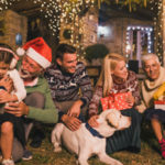 Joyful family holding Christmas presents and petting a dog in front of a decorated house