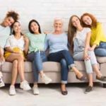 Diverse Women Of Different Age Sitting On Sofa Smiling To Camera Indoor. Support Group Concept