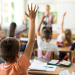 Rear view of little boy and his classmates raising arms to answer teacher's question during the lecture in the classroom.