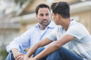 A father sitting with his serious 13 year old teenage son on the curb in front of their house.