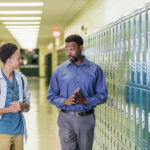 An African-American high school student walking with his teacher or school principal, in the hallway by a row of lockers. They are talking. The teacher has a serious expression on his face, a mature man in his 50s. The male student is an 18 year old teenage boy carrying a backpack.